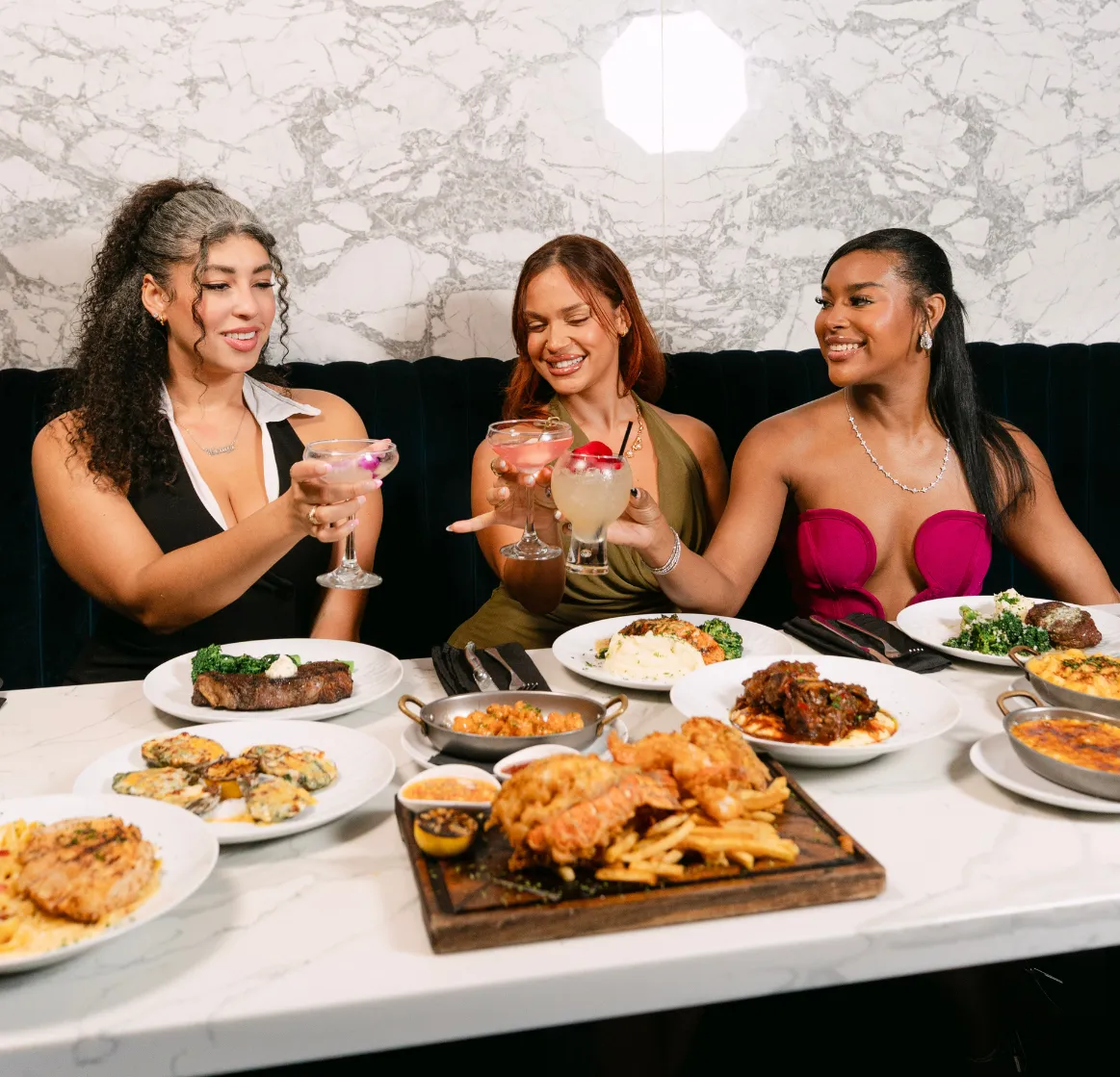 Three women smiling while toasting cocktails over a rich dinner spread at Ciel Restaurant and Lounge.